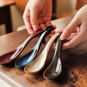 Może przedstawiać: Cztery ceramiczne łyżki w różnych kolorach: bordowym, granatowym, kremowym i nakrapianym brązowym. Każda łyżka ma długi, zakrzywiony uchwyt i głęboką miseczkę. Łyżki są prezentowane na drewnianej powierzchni.