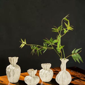 May include: A set of four off-white ceramic vases with textured, ruffled, and organic shapes. One tall vase on the right contains a green artificial bamboo branch with multiple pointed leaves. The vases have narrow necks and bulbous bodies.