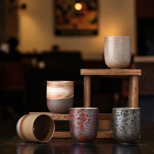 May include: A collection of ceramic cups in various colors and textures. The cups are displayed on a wooden stand and a dark surface. The cups have a rustic, handcrafted appearance, with colors ranging from beige and brown to red and green.