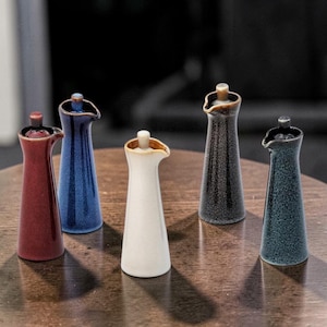 Op de afbeelding: Een verzameling van zes keramische olie- of azijndispensers in verschillende kleuren. De dispensers zijn hoog en slank met een tuit en een kleine stopper. Kleuren zijn rood, blauw, wit, zwart en teal. De dispensers staan op een houten oppervlak.