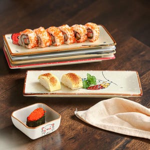 Peut inclure: Présentation de sushi sur des assiettes rectangulaires en céramique. Les assiettes contiennent des rouleaux de sushi avec des œufs de poisson orange, du tamago et un petit plat carré avec un motif de chat. Une serviette beige est également présente.