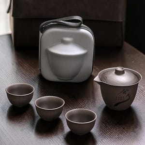 May include: A dark gray tea set with three small cups, a teapot with a lid, and a carrying case. The teapot has a spout and a small design on the side. The cups and teapot are arranged on a dark wooden surface.