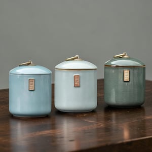 May include: Three ceramic jars in shades of blue and green with matching lids. Each jar has a small, rectangular label and a gold-coloured handle on top. The jars are cylindrical and sit on a dark wooden surface.
