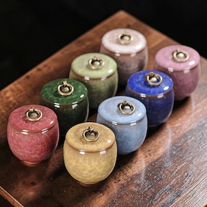 Puede incluir: Una colección de pequeños tarros de cerámica con tapas. Los tarros son de varios colores, incluyendo rojo, verde, azul y amarillo, y tienen un acabado craquelado. Cada tarro tiene un pequeño anillo dorado en la tapa. Están dispuestos sobre una superficie de madera.