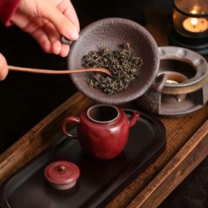 May include: A small, red ceramic teapot with a matching lid sits on a dark tray. A bowl of tea leaves is held above the teapot with a wooden spoon. A candle warmer is in the background, suggesting a tea ceremony.