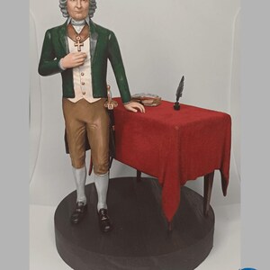 Pode incluir: Estatueta detalhada de uma figura histórica, vestida com um casaco verde, calças castanhas e camisa branca. A figura está ao lado de uma pequena mesa coberta com um pano vermelho, com uma pena e livros. A estatueta está sobre uma base circular castanha escura.
