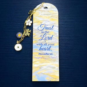 Peut inclure: Un marque-page rectangulaire au motif marbré jaune, blanc et bleu. Il porte le texte bleu Trust in the Lord with all your heart. Proverbs 3:5. Une chaîne dorée est fixée, ornée de breloques en forme de croix, de fleur et de cœur.