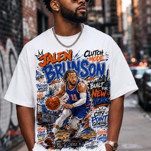 Puede incluir: Una camiseta blanca de manga corta con un estampado gráfico de un jugador de baloncesto con uniforme azul y naranja. El diseño incluye el texto Jalen Brunson, Clutch Mode, Built for New York y City Point General con salpicaduras de pintura.