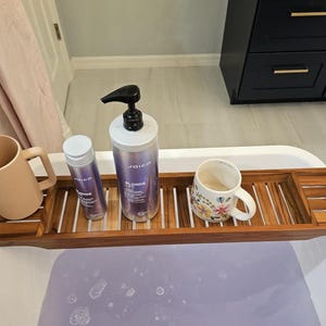May include: A wooden bathtub tray holds a pink mug, a floral mug, and two bottles of Joico Blonde Life shampoo. The tray is positioned over a white bathtub filled with purple water, creating a spa-like atmosphere.
