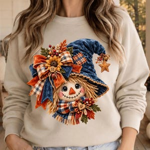 May include: A beige long-sleeve sweatshirt featuring a graphic print of a scarecrow wearing a blue velvet hat. The design includes a sunflower, plaid ribbons, autumn leaves, berries, and a small wooden heart accent on the scarecrow's scarf.