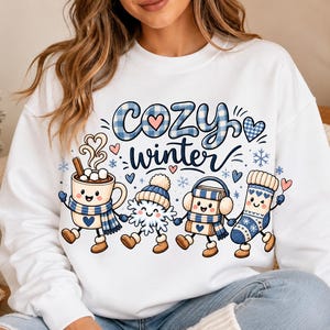 May include: A white long-sleeve sweatshirt featuring a graphic print with the text "cozy winter" in blue plaid and navy script. The design includes four cartoon characters: a mug of cocoa, a snowflake, a book, and a sock, all wearing winter accessories.