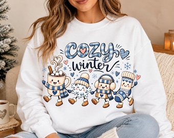 May include: A white long-sleeve sweatshirt featuring a graphic print with the text "cozy winter" in blue plaid and navy script. The design includes four cartoon characters: a mug of cocoa, a snowflake, a book, and a sock, all wearing winter accessories.
