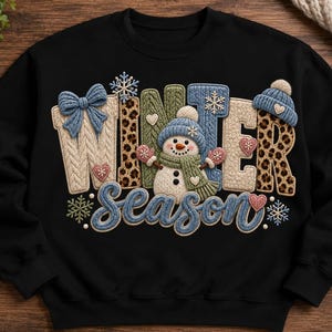 May include: A black long-sleeve sweatshirt featuring a graphic design with the text "WINTER Season". The design includes a central snowman, leopard print lettering, blue knit-textured accents, snowflakes, and small heart shapes in various colors.