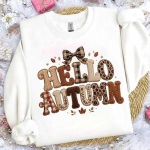 Puede incluir: Una sudadera blanca de manga larga con el texto "HELLO AUTUMN" en letras grandes y decorativas. Las letras tienen varios patrones, incluyendo cuadros, estampado de leopardo y punto trenzado. Un lazo de cuadros se sitúa sobre el texto.