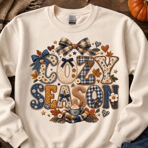 May include: A cream-coloured crewneck sweatshirt featuring a graphic print with the text "COZY SEASON" in large, textured letters. The design includes blue and tan plaid patterns, denim textures, small white flowers, brown hearts, and bows.