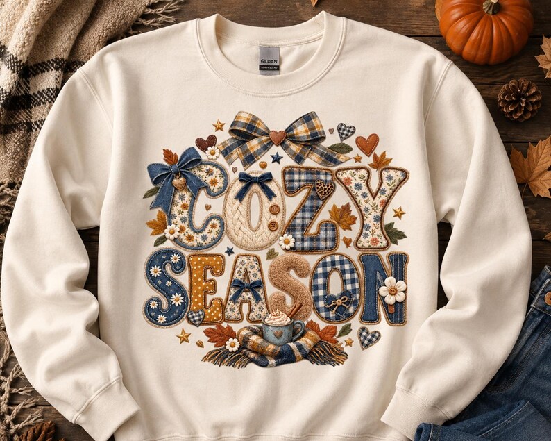 May include: A cream-coloured crewneck sweatshirt featuring a graphic print with the text "COZY SEASON" in large, textured letters. The design includes blue and tan plaid patterns, denim textures, small white flowers, brown hearts, and bows.