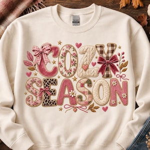 May include: A cream-colored crewneck sweatshirt featuring the text COZY SEASON in large, decorative letters. The letters are filled with various patterns including floral, plaid, leopard print, and cable knit textures, accented with pink bows and hearts.