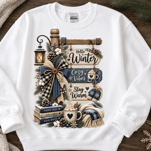 May include: A white crewneck sweatshirt featuring a graphic print of a wooden signpost with three hanging boards that read Hello Winter, Cozy Vibes, and Stay Warm. The design includes a lantern, a plaid bow, pinecones, books, and a mug of cocoa.