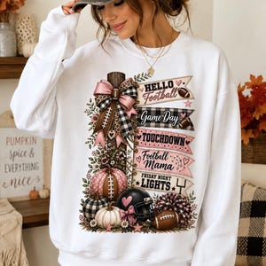 May include: A white long-sleeve sweatshirt featuring a graphic print of a wooden post decorated with pink and black plaid bows, pumpkins, a football helmet, and a pom-pom. The text reads Hello Football, Game Day, TOUCHDOWN, Football Mama, and FRIDAY NIGHT LIGHTS.