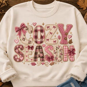 May include: A cream-coloured crewneck sweatshirt featuring the text COZY SEASON in large, decorative letters. The letters are filled with various patterns including plaid, floral, and cable knit textures, accented with pink bows, hearts, and stars.