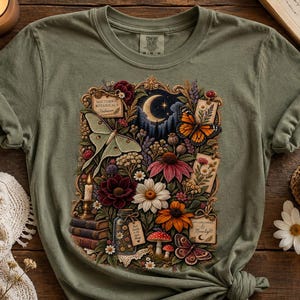 May include: An olive green t-shirt featuring a central graphic print of a crescent moon, a luna moth, a monarch butterfly, various colorful flowers, mushrooms, a stack of books, a lit candle, and small rectangular labels with text.