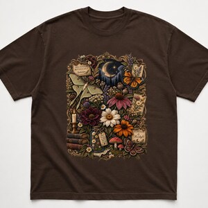 May include: A dark brown short-sleeve t-shirt featuring a central graphic of a moonlit garden scene with a luna moth, monarch butterfly, various colorful flowers, stacked books, a candle, mushrooms, and small rectangular labels with text.