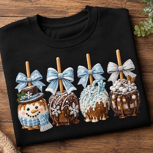 May include: A black crewneck sweatshirt featuring a graphic of four decorated candy apples on sticks. The apples are adorned with light blue bows, white snowflake patterns, chocolate drizzles, and one apple is styled as a snowman with a hat.