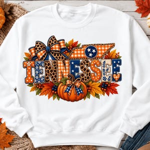 May include: A white long-sleeve sweatshirt featuring a graphic print of the word TENNESSEE in block letters with various patterns including leopard print, plaid, and polka dots. The design includes a pumpkin, autumn leaves, and a bow.