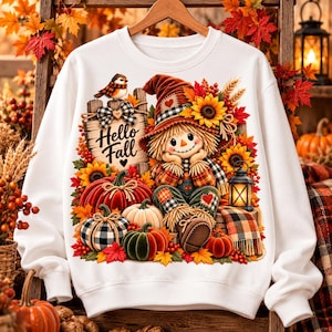 May include: A white long-sleeve sweatshirt featuring a graphic of a scarecrow wearing a hat, surrounded by sunflowers, pumpkins, and a sign that reads Hello Fall. A small bird sits on the sign, and a lantern is placed next to the scarecrow.