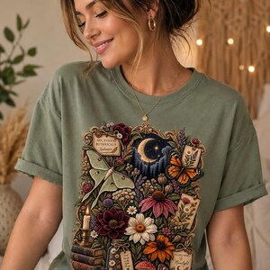 May include: An olive green t-shirt featuring a detailed graphic print of nocturnal botanicals, including a crescent moon, a moth, a butterfly, various colorful flowers, a lit candle, stacked books, and small labels with text.