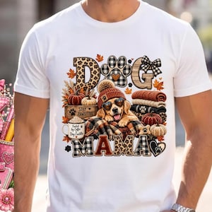 Puede incluir: Una camiseta blanca de manga corta con un gr&aacute;fico de un golden retriever con gorro y gafas de sol en forma de coraz&oacute;n. El dise&ntilde;o incluye el texto DOG MAMA, una taza de caf&eacute; con la inscripci&oacute;n Fueled by Coffee & Dog Cuddles, calabazas y hojas.