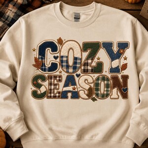 May include: A cream-coloured crewneck sweatshirt featuring the text COZY SEASON in large, block-style letters. Each letter is filled with various plaid and solid fabric patterns in shades of blue, green, brown, and white, accented by small stars, hearts, and leaves.
