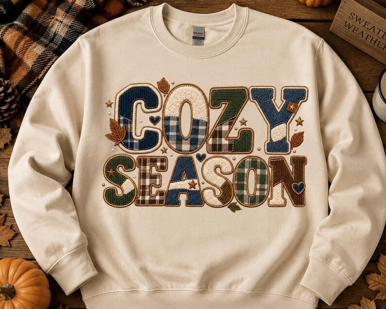 May include: A cream-coloured crewneck sweatshirt featuring the text COZY SEASON in large, block-style letters. Each letter is filled with various plaid and solid fabric patterns in shades of blue, green, brown, and white, accented by small stars, hearts, and leaves.
