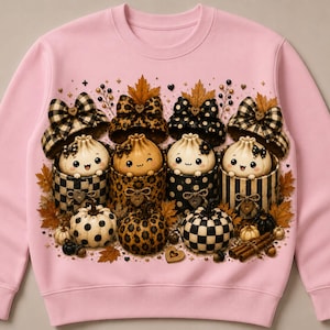 May include: A light pink crewneck sweatshirt featuring a graphic print of four cartoon characters resembling dumplings, each sitting in a patterned container topped with a bow. The design includes autumn leaves, small pumpkins, and cinnamon sticks.
