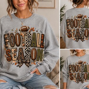 May include: A light gray crewneck sweatshirt featuring the text "FOOTBALL SEASON" in large, blocky letters. The letters are decorated with various patterns including leopard print, plaid, and solid olive green. A brown football and a plaid bow are included.