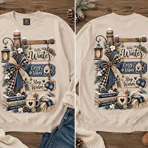 May include: A beige crewneck sweatshirt featuring a graphic print of a wooden signpost with three hanging signs that read Hello Winter, Cozy Vibes, and Stay Warm. The design includes a lantern, a plaid bow, pinecones, books, and a mug of cocoa.