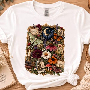 May include: A white short-sleeve t-shirt featuring a central graphic print of a gold ornate frame containing a crescent moon, a pale green moth, a monarch butterfly, various colorful flowers, mushrooms, stacked books, and a candle.