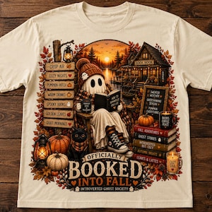 May include: A cream-coloured t-shirt featuring a graphic of a ghost reading a book in a cosy autumn setting. The design includes a black cat, pumpkins, lanterns, a stack of books, and text that reads "OFFICIALLY BOOKED INTO FALL" and "INTROVERTED GHOST SOCIETY".