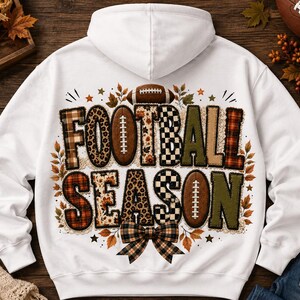May include: A white hooded sweatshirt featuring a graphic on the back with the text FOOTBALL SEASON. The letters are filled with various patterns including leopard print, plaid, and checkered designs, accented by footballs and autumn leaves.