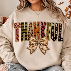 May include: A beige crewneck sweatshirt featuring the word THANKFUL in large, multi-patterned letters. Each letter displays a unique design, including checkered, striped, leopard, pumpkin, and ghost prints. A large, floral-patterned bow sits below.