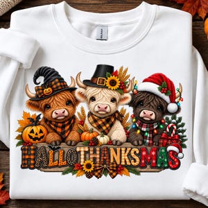 Op de afbeelding: Een wit sweatshirt met lange mouwen en een afbeelding van drie Highland-koeien met seizoensgebonden hoeden: een heksenhoed, een pelgrimshoed en een kerstmuts. Daaronder staat de tekst HALLOTHANKSMAS op een houten bord afgebeeld.