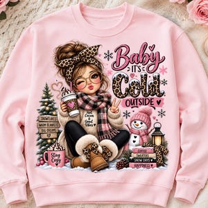 Op de afbeelding: Een lichtroze sweatshirt met een afbeelding van een persoon met een luipaardprint hoofdband, bril en sjaal, die een beker vasthoudt. De tekst luidt Baby It's Cold Outside, Hot Cocoa & Good Vibes, Stay Cozy, Snowflakes, Warm Blankets, Big Dreams, Coffee, Sweaters, Snow Days, Happiness.