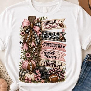May include: A white t-shirt featuring a graphic print of a wooden post decorated with a pink and black plaid bow, pumpkins, a football helmet, and a pom-pom. The text reads: Hello Football, Game Day, TOUCHDOWN, Football Mama, and FRIDAY NIGHT LIGHTS.