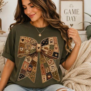 Könnte beinhalten: Ein olivgrünes T-Shirt mit einer großen, mittigen Grafik einer Schleife, die aus einem Patchwork verschiedener brauner, beiger und cremefarbener Stoffmuster besteht. Das Schleifendesign enthält kleine Abbildungen von Kaffeetassen, Herzen und Sternen.