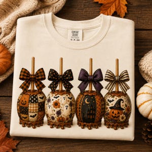 May include: A cream-colored t-shirt with a graphic print of four candy apples, each topped with a unique patterned bow. The designs feature pumpkins, ghosts, bats, a haunted house, and a witch hat. The shirt has a Comfort Colors brand label.