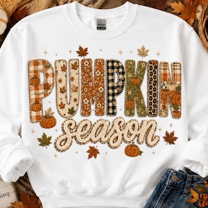 May include: A white crewneck sweatshirt featuring the text PUMPKIN SEASON in large, multi-patterned letters. The letters display various autumn-themed designs including orange plaid, floral prints, black and white buffalo check, and leopard spots.