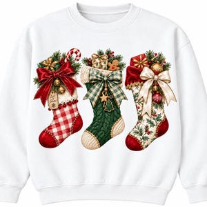 Puede incluir: Una sudadera blanca de cuello redondo con un gráfico de tres calcetines navideños. El calcetín izquierdo es de cuadros rojos y blancos con un lazo rojo grande y una etiqueta que dice Let it Snow. El central es de punto verde con un lazo de cuadros.