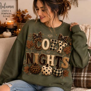 May include: An olive green crewneck sweatshirt featuring the text COZY NIGHTS in a mix of leopard, plaid, and polka dot patterns. The design includes two leopard print bows, small hearts, and three pumpkins with matching patterns and glittery leaves.
