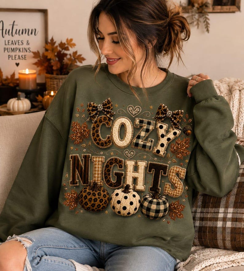 May include: An olive green crewneck sweatshirt featuring the text COZY NIGHTS in a mix of leopard, plaid, and polka dot patterns. The design includes two leopard print bows, small hearts, and three pumpkins with matching patterns and glittery leaves.