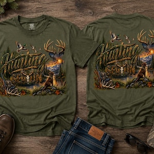 May include: An olive green t-shirt featuring a graphic print with the text Hunting Season. The design includes a brown dog, a deer skull, antlers, flying ducks, a rifle, and a mountain landscape scene with a sunset over a lake and forest.